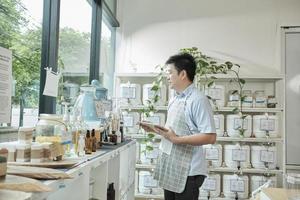 Asian male shopkeeper checks stock of natural organic products at window display in refill store, zero-waste and plastic-free grocery, eco environment-friendly, sustainable lifestyles, reusable shop. photo