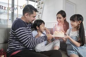 Happy Asian Thai family, young son is surprised with birthday cake, gift, blows out candle, and celebrates party with parents together in living room, wellbeing domestic home event lifestyle. photo