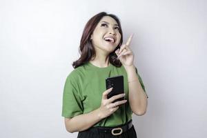 Smiling beautiful Asian woman pointing finger upward to empty space while holding her phone isolated white background photo