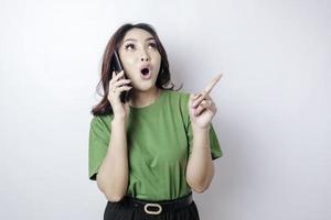 Shocked Asian woman wearing a green t-shirt pointing at the copy space upside her holding her phone, isolated by a white background photo