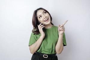 Smiling beautiful Asian woman pointing finger upward to empty space while holding her phone isolated white background photo