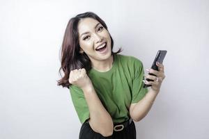 una joven asiática con una expresión feliz y exitosa que usa una camiseta verde y sostiene un teléfono inteligente aislado de fondo blanco foto