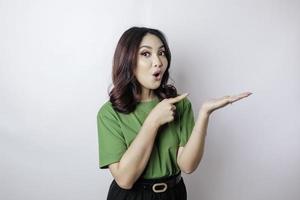 Shocked Asian woman wearing green t-shirt pointing at the copy space beside her, isolated by white background photo