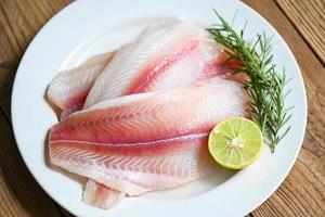 filete de pescado en un plato blanco con ingredientes para cocinar, filete de pescado pangasius crudo fresco con hierbas y especias, lima limón y romero, pescado de carnita tilapia bagre rayado foto