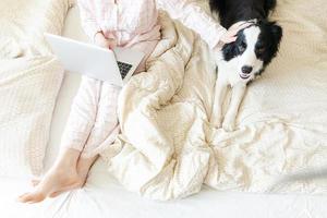oficina móvil en casa. mujer joven en pijama sentada en la cama con un perro mascota trabajando en una computadora portátil en casa. chica de estilo de vida estudiando en el interior. concepto de cuarentena empresarial independiente. foto