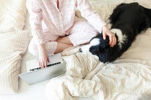 oficina móvil en casa. mujer joven en pijama sentada en la cama con un perro mascota trabajando en una computadora portátil en casa. chica de estilo de vida estudiando en el interior. concepto de cuarentena empresarial independiente. foto