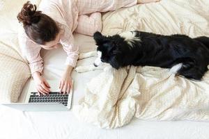 oficina móvil en casa. mujer joven en pijama sentada en la cama con un perro mascota trabajando en una computadora portátil en casa. chica de estilo de vida estudiando en el interior. concepto de cuarentena empresarial independiente. foto