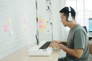 Asian man wearing white headphones play piano and sing a song and learning online with mobile phone and compose writing song or record sound with a microphone and use computer recording music program photo