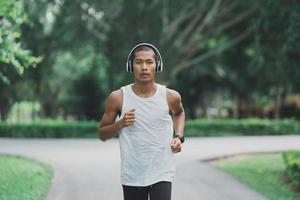 Asian man sportsman fit body wearing white headphone and smart watch while running or jogging in the garden have more tree and clean air, exercising at park. Sport healthy running concept. photo