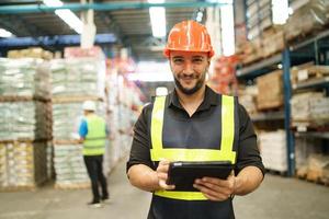 Professional manager man employee using tablet check stock working at warehouse. Worker wearing high visibility clothing and a hard hat, helmet and checking and count up goods or boxes for delivery. photo