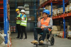 Manager handicapped in wheelchair man employee using tablet check stock work at warehouse. Worker wearing high visibility clothing and hard hat, helmet and checking count up goods boxes for delivery. photo