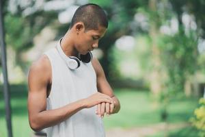 Asian man sportsman fit body looking and touch screen to smart watch and wearing white headphone while resting or before jogging, exercising at park. Sport healthy running concept. photo