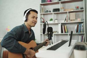 Asian man wearing white headphones play guitar and sing a song and learning online with mobile phone and compose writing song or record sound with a microphone and use computer recording music photo