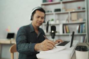 Asian man wearing white headphones play piano and sing a song and learning online with mobile phone and compose writing song or record sound with a microphone and use computer recording music program photo