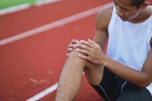 Athletes sport man runner wearing white sportswear to sitting feeling pain in his leg or knee after practicing on a running track at a stadium, copy space. Runner sport injury concept. photo