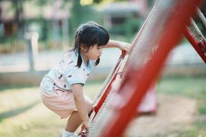 linda niña asiática sonríe jugar en la escuela o en el jardín de infantes o en el patio de recreo. Actividad de verano saludable para niños. niña asiática escalando al aire libre en el patio de recreo. niño jugando en el patio de recreo al aire libre. foto