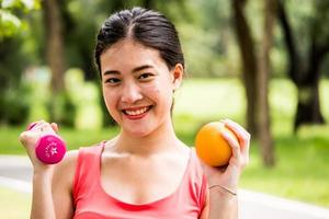 Portrait of attractive woman is holding dumbbell and orange in the park. photo