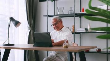 An elderly man reading news with the tablet on the desk at home. An old Asian man is searching for information on the Internet while sitting at a table. photo