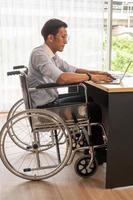 Asian businessman sitting on wheelchair and typing on laptop at the office. photo