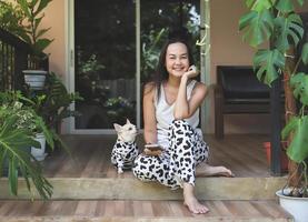 Asian woman and her chihuahua dog wearing dalmatian pattern costume sitting on balcony with house plants, smiling and looking at camera. photo