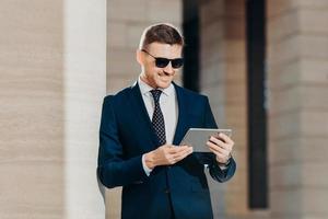 People, style, business and technology concept. Horizontal shot of attractive young businessman reads important information on tablet computer, connected to wifi, wears trendy shades and suit photo