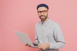 Talented male journalist with appealing look, holds modern laptop computer, works on new article, dressed in fashionable clothes, models against pink background, connected to wireless internet photo