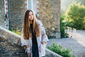 Portrait of beautiful young female model wears warm mantlet, keeps hands in pocket, poses against ancient landmarks, enjoys wonderful nature and calm atmosphere, admires archtectural builduings photo