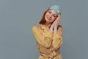 Beautiful relaxed woman tilts head on palms pressed together, looks at camera with satisfaction, enjoys late wake up and having enough sleep, wears pajamas and blindfold, isolated on grey wall photo