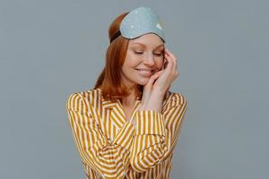 Sweet dreams and good rest concept. Pretty redhead woman turns head aside, keeps gaze down and touches cheek gently, wears striped pajama, blindfold, smiles positively, isolated over grey wall photo