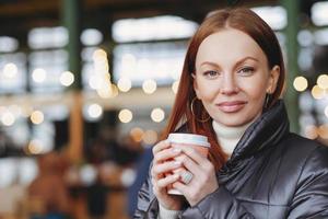 toma exterior de una modelo femenina atractiva y satisfecha con piel sana, cabello castaño, toma café para llevar, disfruta de una buena bebida, vestida con chaqueta, espera a alguien, toma un descanso para tomar café, mira la cámara foto