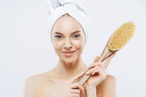 Pretty young woman with healthy skin, takes care of personal hygiene, holds bath brush, wrapped towel on head, stands shirtless indoor, white background. Women, skin care, freshness concept. photo