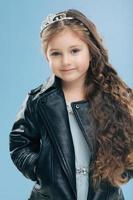 Isolated shot of beautiful Caucasian small child has blue eyes, long dark hair, keeps hands in pockets of leather jacket, isolated over blue background. Children, style, childhood, fashion concept photo