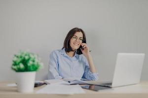 Beautiful female recruiter makes job offer to somene via cell phone, checks curriculum vitae on laptop computer, expalins detailes and pluses of work, surrounded with flower and personal diary photo