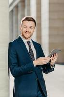 People, work and technology concept. Handsome male economist searches information online on digital tablet, prepares for meeting with staff, works on financial report, dressed in formal clothing photo