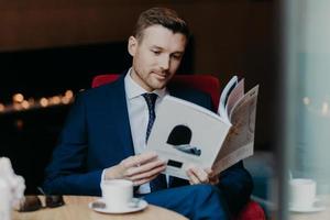 Concentrated businessman looks attentively in menu while sits in cozy restaurant, drinks coffe, dressed in formal suit, has appealing look, reads some information, compares prices in cafeteria photo