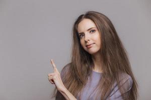 Horizontal shot of beautiful woman with long straight hair, pleasant appearance, dressed in casual sweater, poses against grey background with blank grey wall for your advertising content or promotion photo