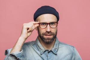 Portrait of pleasant looking satisfied bearded man with pleasant appearance looks confidently through spectacles, wears fashionable clothing, poses against pink studio background. Facial expressions photo