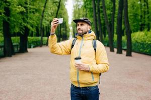 Outdoor portrait of handsome guy with thick beard wearing yellow anorak and jeans holding rucksack, coffee and smartphone making selfie against green trees. Pleased tourist having rest in park photo