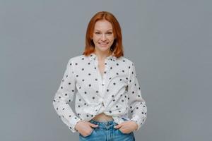 Fashionable positive woman wears polka dot shirt and jeans, keeps hands in pockets, stands self assured against grey background, has happy casual talk with interlocutor. Emotions, fashion concept photo