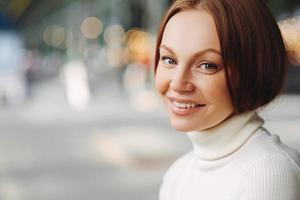 Sideways shot of pleasant looking woman with make up, tender smile, dressed in white turtleneck sweater, stands against blurred background with copy space for your advertisement, has healthy skin photo