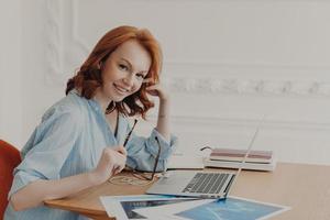 Satisfied businesswoman with foxy hair, checks accounting documentation in online database, works on modern laptop indoors, poses at workplace, surrounded with papers, thinks about creative ideas photo