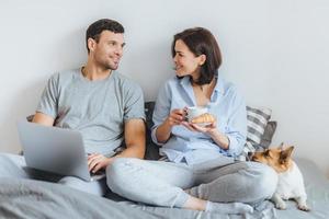 Portrait of lovely couple sit together on bed, use laptop computer, have pleasant conversation between each other, do shopping online. Jack russell terrier lies near hosts in bedroom photo