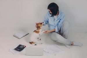 Brunette woman dressed casually, studies income report, sits on floor with laptop computer, paper documents, calculator, notepad, has fun with her favourite pet, isolated over white background photo