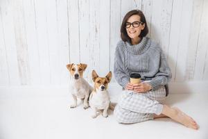 hermosa mujer morena se sienta en el suelo con sus dos perros favoritos, vestida de manera informal, bebe café para llevar. la mujer complacida disfruta de un ambiente doméstico tranquilo. personas y animales, buenas relaciones foto