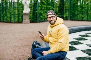 Horizontal portrait of bearded hipster male looking aside with delightful expression while sitting near fountain making photos of beautiful nature. Stylish man resting outdoors holding cell phone