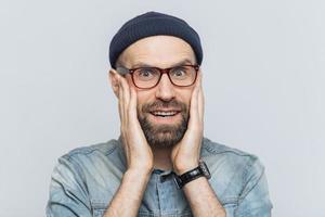 Indoor shot of excited bearded man with blue eyes and cheerful expression, keeps hands on cheeks, dressed in fashionable clothing, isolated over white background. People, facial expressions concept photo