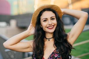 Happy brunette female with pleasant smile wearing hat keeping hand behind head posing outdoors. Pretty woman with happy expression relaxing at big city during her summer holidays. People, emotions photo