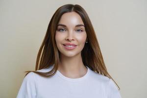 Headshot of beautiful dark haired young European woman looks with tender expression, has friendly attitude, talks casually with friends, wears casual white t shirt, isolated over beige background photo
