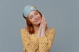 Adorable freckled red haired woman keeps eyes closed, hands together near freckled face, wears sleepmask and casual pajama, isolated over grey background, has pleasant dreams during afternoon nap. photo