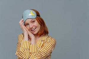 Morning and wake up. Cute cheerful redhead young European woman wears blindfold and nightwear, looks gladfully at camera, isolated over grey studio wall with empty space, awakes in good mood. photo